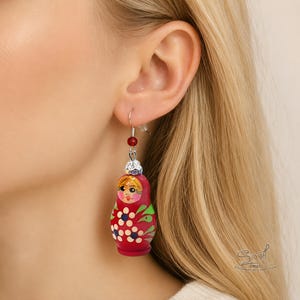Close-up of a blonde woman wearing a hand-painted red Matryoshka doll earring with floral details, silver hook, and red bead accent. Large Russian nesting doll dangle earring shown on ear to display size and intricate artisan painting
