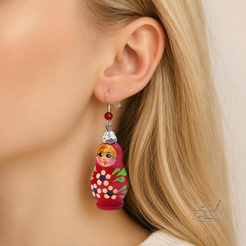 Close-up of a blonde woman wearing a hand-painted red Matryoshka doll earring with floral details, silver hook, and red bead accent. Large Russian nesting doll dangle earring shown on ear to display size and intricate artisan painting