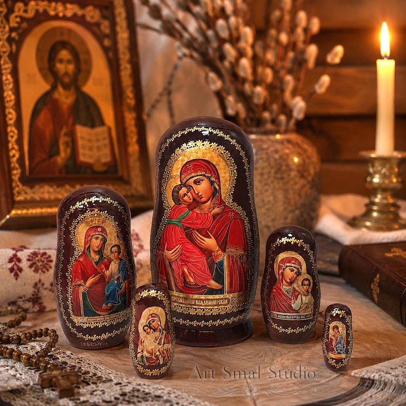 May include: A set of five dark brown nesting dolls featuring hand-painted icons of the Virgin Mary and Child in vibrant red and blue robes. Each doll is decorated with intricate gold leaf detailing along the edges and base, showcasing traditional religious artistry with a glossy finish.