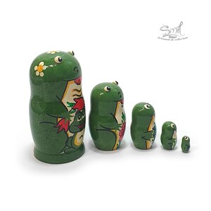 Shown from a side angle, the five green frog nesting dolls display their rounded wooden shapes, glossy finish, floral decorations and colorful painted frog artwork