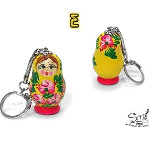 May include: Two yellow Matryoshka doll keychains. The dolls have a red base and are decorated with pink flowers and green leaves. The keychains have silver-colored metal rings and attachments. The text "E" is in a yellow box in the upper left corner.