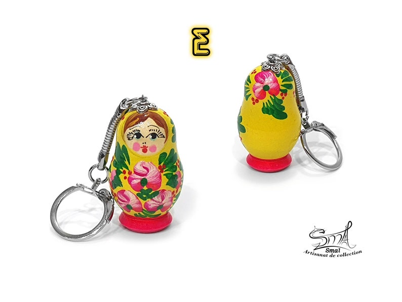 May include: Two yellow Matryoshka doll keychains. The dolls have a red base and are decorated with pink flowers and green leaves. The keychains have silver-colored metal rings and attachments. The text "E" is in a yellow box in the upper left corner.