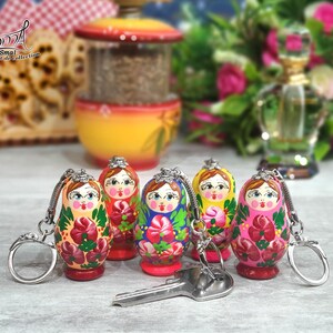 May include: A collection of hand-painted Russian nesting doll keychains. Each doll features a unique floral design in vibrant colors like red, pink, and green. The keychains are attached to silver-colored metal rings. A key rests in front of the dolls.
