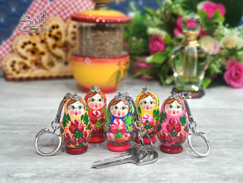 May include: A collection of hand-painted Russian nesting doll keychains. Each doll features a unique floral design in vibrant colors like red, pink, and green. The keychains are attached to silver-colored metal rings. A key rests in front of the dolls.