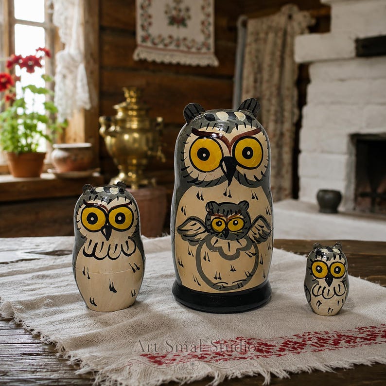 Hand-painted owl nesting dolls displayed on a traditional woven cloth in a rustic Russian cottage interior, with a wooden log wall, brass samovar, lace curtains, potted flowers and a white masonry stove in the background