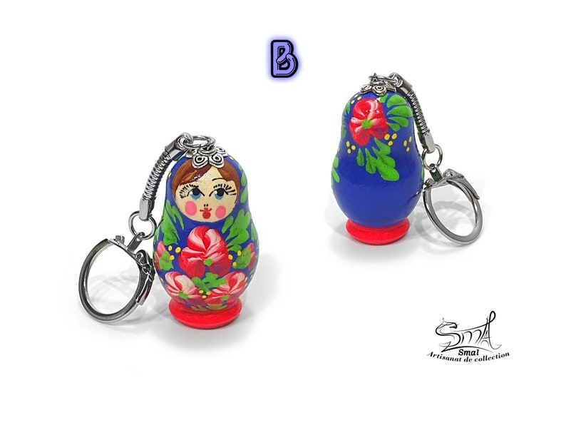 May include: Two Russian nesting doll keychains. The dolls are painted blue with red and green floral designs. Each doll has a silver-colored keychain ring attached to the top. The base of each doll is red. The text "B" is in the upper left corner.