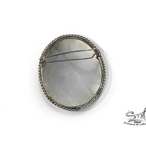 May include: A circular brooch with a silver-coloured twisted rope-style edge. The reverse side of the brooch has a pin clasp. The brooch is set against a white backdrop. A vintage accessory.