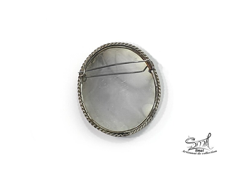 May include: A circular brooch with a silver-coloured twisted rope-style edge. The reverse side of the brooch has a pin clasp. The brooch is set against a white backdrop. A vintage accessory.