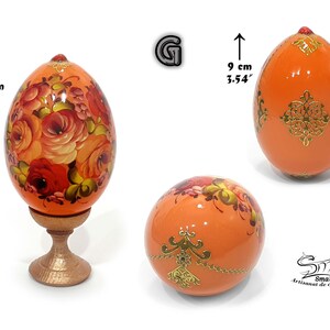 Œuf de Pâques en teinte orange clair nacré, orné d’un décor floral peint à la main inspiré de l’art russe, sublimé par des motifs dorés délicats, avec socle inclus, hauteur totale 12,5 cm, hauteur de l’œuf 9 cm