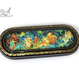 Può includere: Una scatola di legno laccata nera di forma ovale con un bordo dorato. Il coperchio è decorato con una scena di arte popolare colorata che rappresenta una carrozza trainata da cavalli, figure e un villaggio.