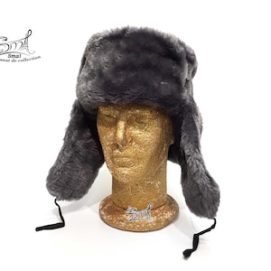 Op de afbeelding: Een grijze imitatiebont ushanka-muts met oorflappen en een zwart trekkoord. De muts zit op een mannequinhoofd.