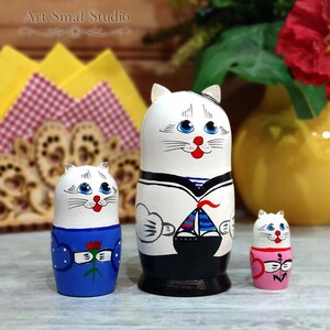 Hand-painted Russian cat sailor matryoshka dolls displayed as a set of three wooden nesting dolls, photographed on a tabletop with a yellow vase and decorative napkin holder in the background