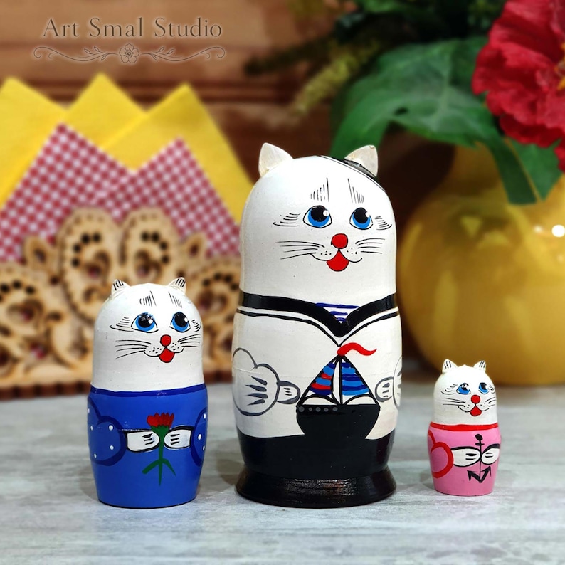 Hand-painted Russian cat sailor matryoshka dolls displayed as a set of three wooden nesting dolls, photographed on a tabletop with a yellow vase and decorative napkin holder in the background