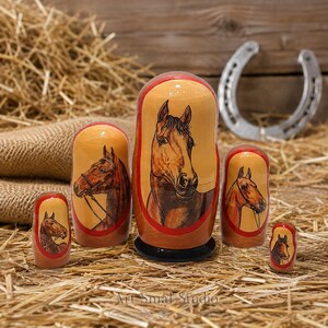 Hand painted wooden horse nesting dolls displayed on straw with a rustic horseshoe backdrop, featuring five detailed equestrian portraits and traditional Russian folk art