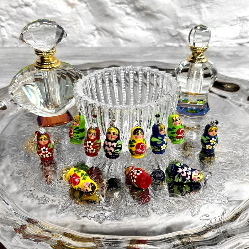 Collection of handmade Matryoshka earrings in multiple colors and floral designs, miniature Russian nesting doll jewelry displayed on vintage silver tray with crystal glass decor, colorful wooden babushka earrings by ArtSmalStudio, unique artisan folk art gift for women