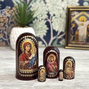 May include: A set of six Russian nesting dolls, each featuring religious iconography. The dolls are painted in deep red with gold accents and depict figures with halos. The largest doll is approximately 6 inches tall.