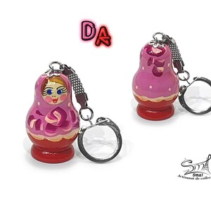 May include: A pink and red wooden nesting doll keychain with a painted face and floral design. The keychain is attached to a silver metal ring.