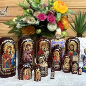 May include: A collection of maroon nesting dolls, each adorned with religious iconography in gold and vibrant colors. The dolls vary in size, with the largest standing prominently. A small angel figurine and a bouquet of flowers are also present.