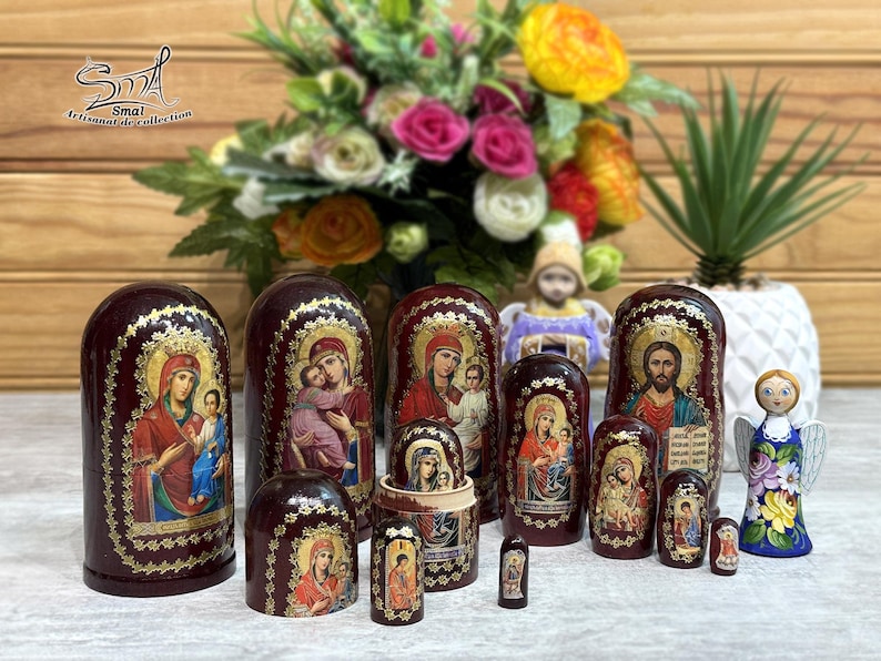 May include: A collection of maroon nesting dolls, each adorned with religious iconography in gold and vibrant colors. The dolls vary in size, with the largest standing prominently. A small angel figurine and a bouquet of flowers are also present.