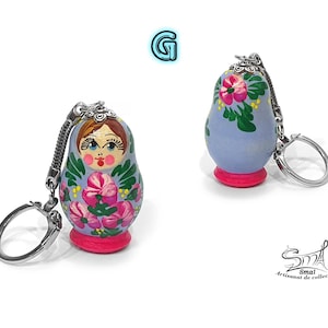 May include: Two Matryoshka doll keychains. The dolls are painted with floral designs in pink, green, and white on a light blue background. Each doll has a pink base and a silver keychain attachment. The image includes the text "Smai Artisanat de collection".