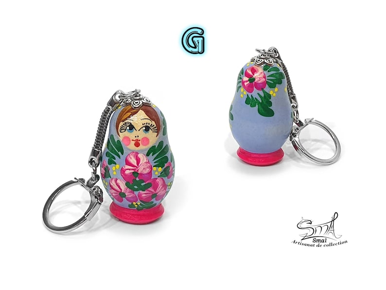 May include: Two Matryoshka doll keychains. The dolls are painted with floral designs in pink, green, and white on a light blue background. Each doll has a pink base and a silver keychain attachment. The image includes the text "Smai Artisanat de collection".