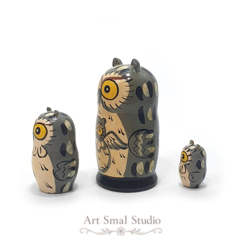 Side view of a three-piece hand-painted owl matryoshka nesting doll set, showing detailed gray, cream and dark blue folk-art patterns, wings, feathers and yellow eyes on a clean white background