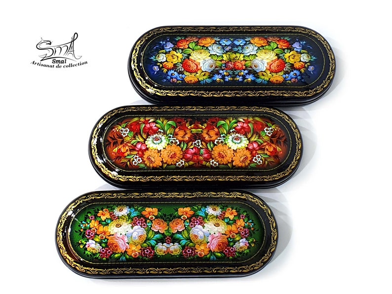 Puede incluir: Tres cajas ovaladas negras con intrincados bordes dorados y dise&ntilde;os florales pintados en colores vibrantes. Las cajas est&aacute;n apiladas una encima de la otra, con la caja superior con un fondo azul, la caja del medio con un fondo naranja y la caja inferior con un fondo verde.