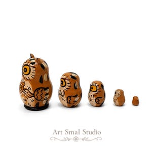 Viewed from the side, this five-piece owl nesting doll set reveals the detailed feathers, wings and warm brown coloring of each graduated wooden figure, from the largest owl to the tiny inner doll, presented against a crisp white background