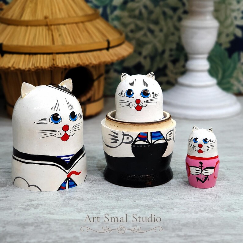 A cozy tabletop scene showcases three hand-painted Russian cat sailor matryoshkas, with the doll opened to reveal the smaller cat inside. A wicker basket, white decorative vase and green plants create a warm home setting in the background