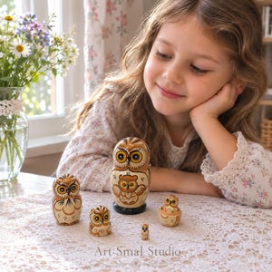 Matriochka hiboux russe en bois à 5 pièces, présentée sur une table en dentelle avec une jeune fille qui la regarde, dans un intérieur lumineux avec des fleurs et une fenêtre