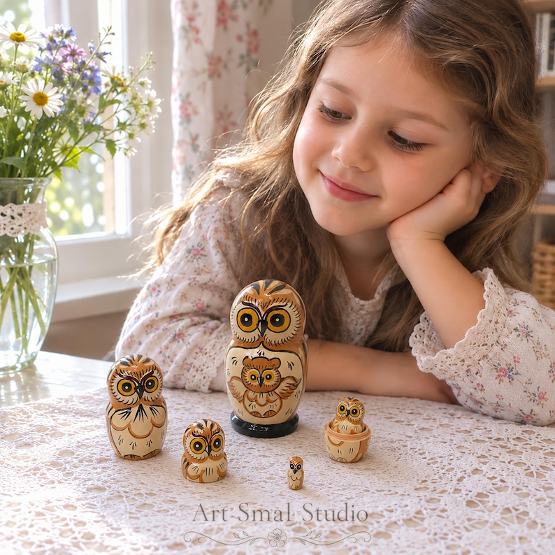 Matriochka hiboux russe en bois à 5 pièces, présentée sur une table en dentelle avec une jeune fille qui la regarde, dans un intérieur lumineux avec des fleurs et une fenêtre