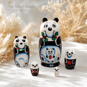 Panda matryoshka nesting dolls shown in an unassembled arrangement, with all five Russian nesting dolls separated and displayed individually on a white tabletop. The scene has a softly blurred background of dried flowers and warm beige foliage, creating an elegant product setting