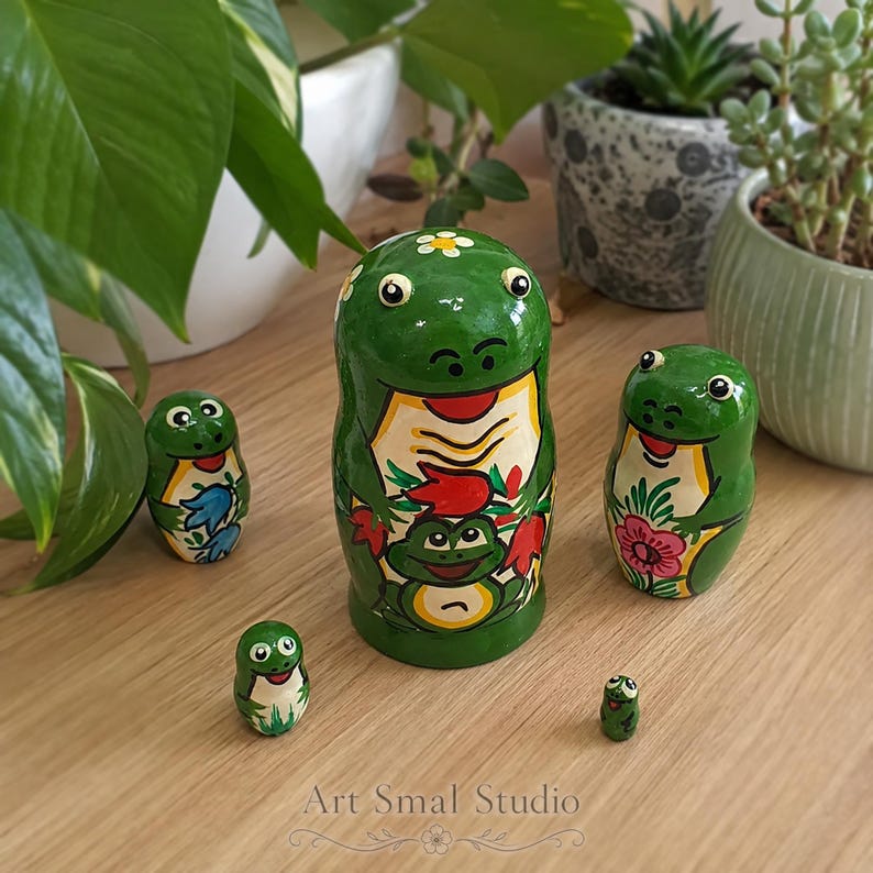 A charming frog matryoshka collection is arranged on a light wooden table among lush houseplants, showcasing five hand-painted green nesting dolls with colorful floral and frog motifs
