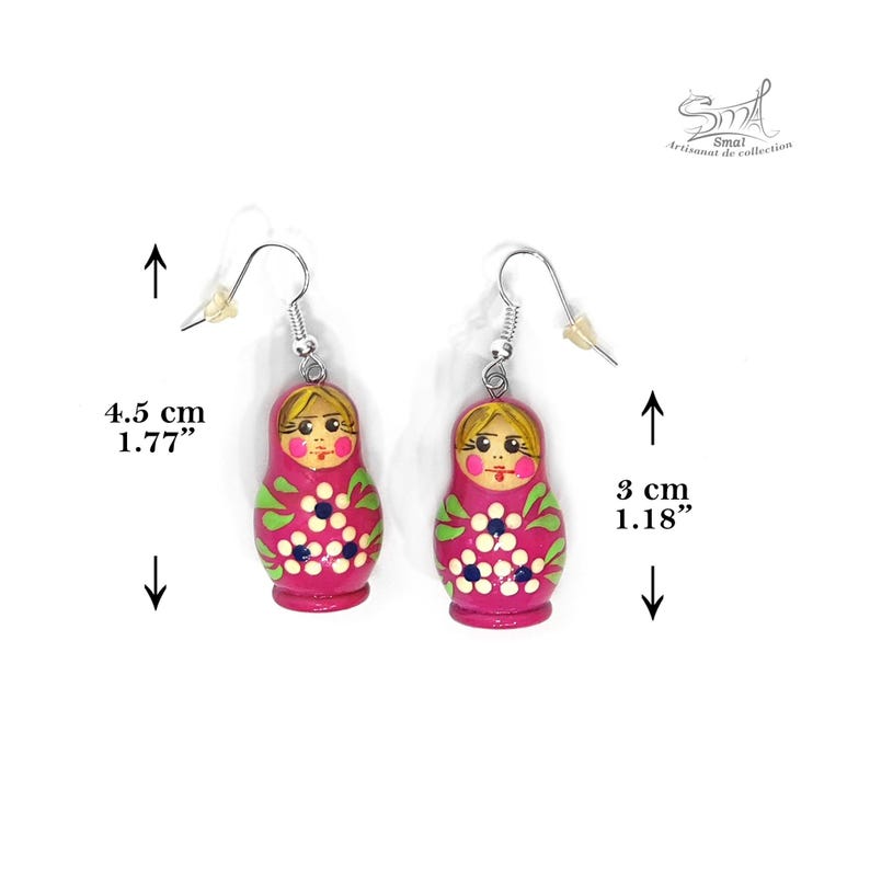 Boucles d'oreilles poupées russes roses avec crochets argentés. Chaque poupée mesure 4,5 cm de haut et présente un visage peint, des feuilles vertes et des fleurs blanches. Les boucles mesurent 3 cm de large.