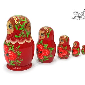May include: A set of six red nesting dolls, each decreasing in size. The dolls feature painted faces, floral designs, and strawberry motifs. The largest doll is approximately 15 cm tall. The dolls are arranged in a row against a white background.