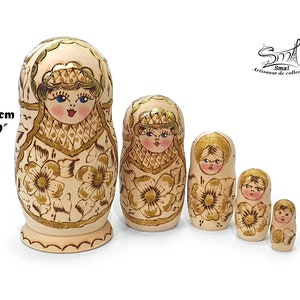 May include: Set of five traditional Russian nesting dolls, or matryoshka dolls. The dolls are painted in a cream colour with gold floral designs and glitter accents. The largest doll measures 15.5 cm tall.