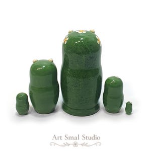 The back view of this five-piece frog nesting doll set shows the smooth green wooden bodies, glossy finish, tiny raised eyes, and floral decorations on the largest doll