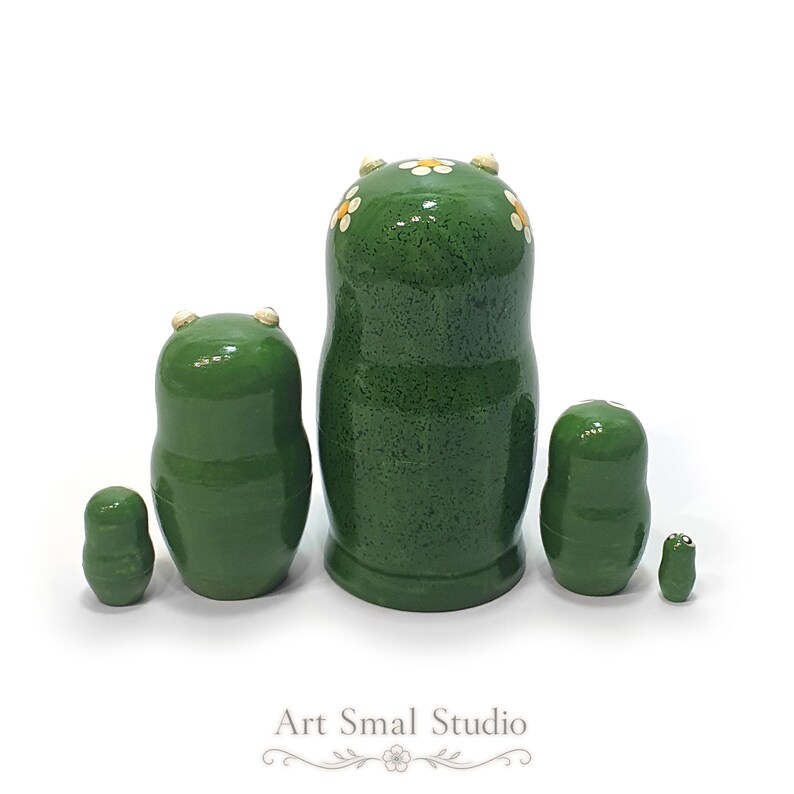 The back view of this five-piece frog nesting doll set shows the smooth green wooden bodies, glossy finish, tiny raised eyes, and floral decorations on the largest doll