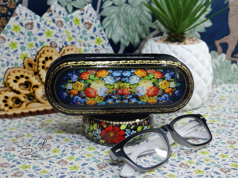 Puede incluir: Un estuche para gafas ovalado de laca negra con un dise&ntilde;o floral. El estuche est&aacute; decorado con una pintura tradicional de arte popular ruso de flores y hojas coloridas. El estuche est&aacute; sentado en un peque&ntilde;o soporte con un dise&ntilde;o floral similar. Un par de gafas de lectura negras y blancas est&aacute;n tiradas sobre una superficie estampada.