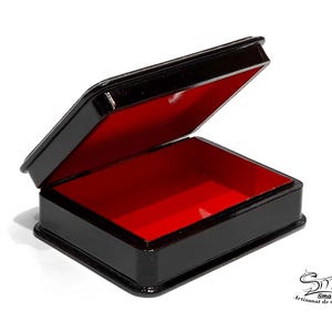 May include: A small, rectangular black box with a hinged lid, open to reveal a vibrant red interior. The box is glossy and appears to be made of a lacquered material. The box is on a white background. The box is labeled with the text "Smal Artisanat de collection".