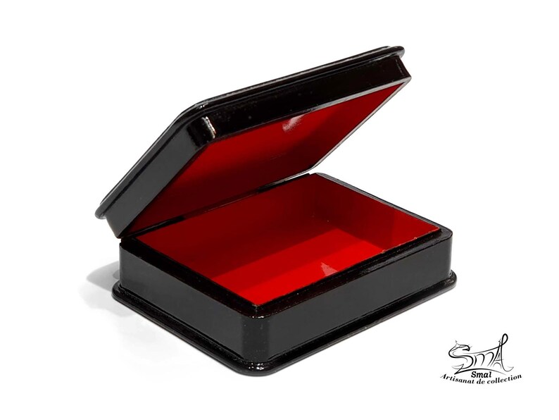 May include: A small, rectangular black box with a hinged lid, open to reveal a vibrant red interior. The box is glossy and appears to be made of a lacquered material. The box is on a white background. The box is labeled with the text "Smal Artisanat de collection".