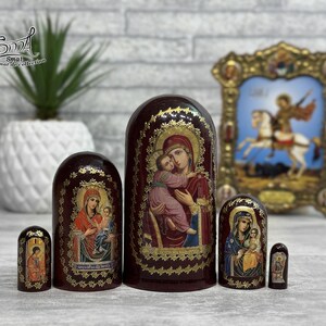 May include: A set of six Russian nesting dolls, each with a dark red base and gold accents. The dolls feature religious iconography, including images of the Virgin Mary and Jesus. A framed religious icon is in the background.