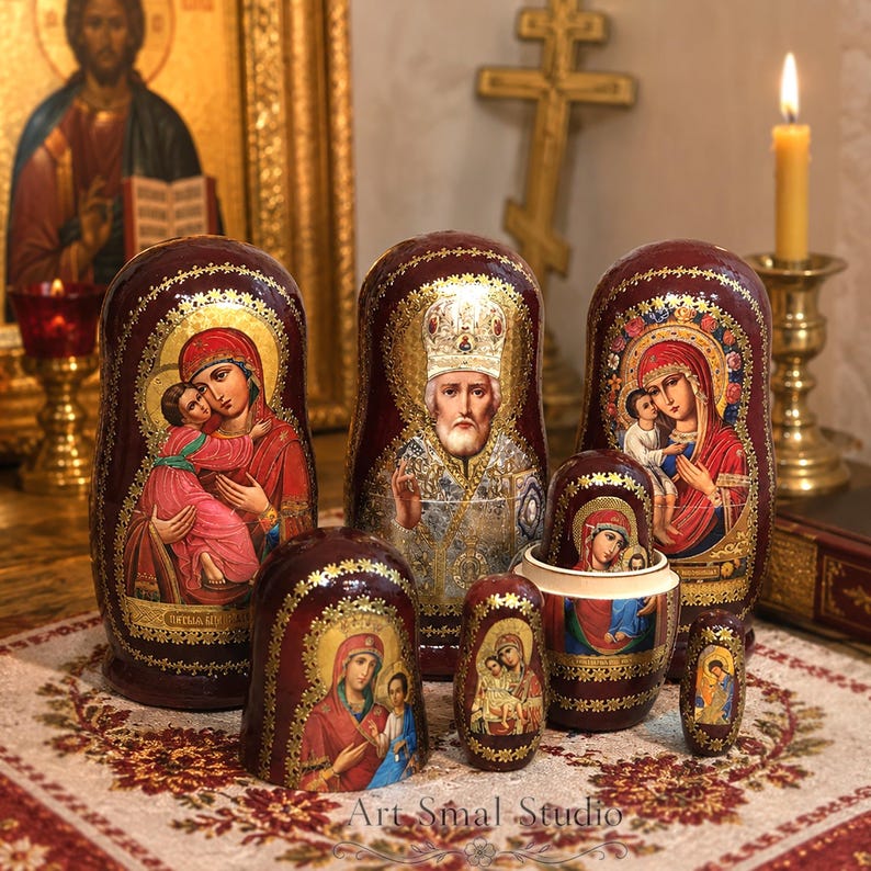 May include: A set of six nesting dolls with a deep red finish and gold leaf detailing. Each doll features a hand-painted religious icon, including depictions of the Virgin Mary with the Christ child and Saint Nicholas, with Art Smal Studio text.