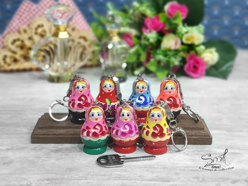 May include: A set of seven colorful wooden nesting doll keychains. Each doll is painted with a different design, including flowers, geometric patterns, and traditional Russian folk art. The keychains are attached to silver rings.