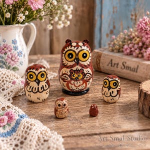 Charming owl nesting doll set with five hand-painted wooden owls in warm brown, cream and golden tones, including one of the world’s smallest matryoshka dolls. Shown on a rustic wooden table with pink and white flowers, a floral pitcher, blue wooden backdrop and Art Smal book