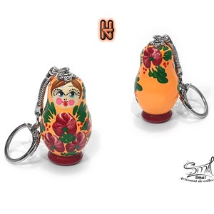 May include: Orange Matryoshka doll keychain with a floral design in red and green. The doll has a silver keychain and a red base. The doll's face is painted with rosy cheeks and a stern expression. The text "Smai Artisanat de collection" is in the bottom right corner.