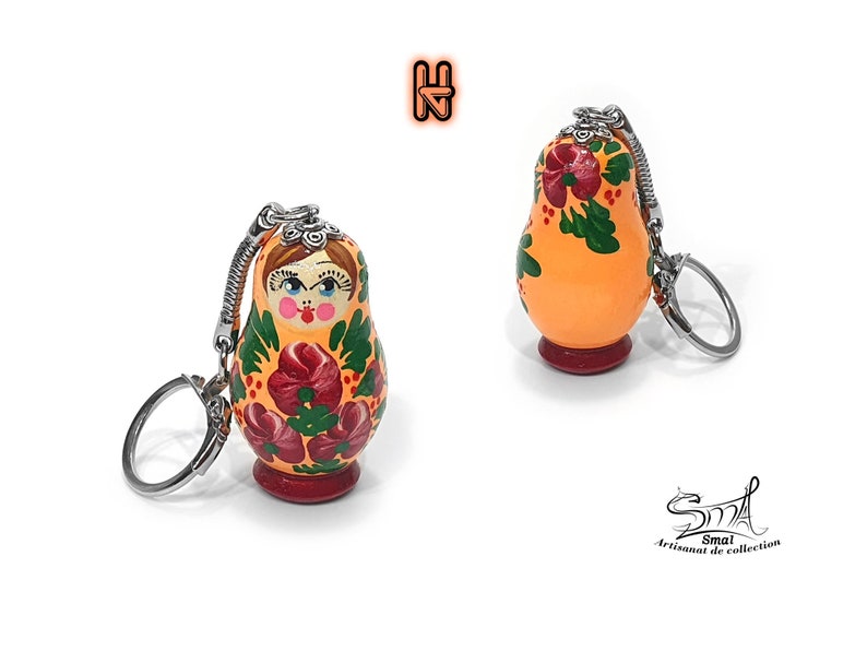 May include: Orange Matryoshka doll keychain with a floral design in red and green. The doll has a silver keychain and a red base. The doll's face is painted with rosy cheeks and a stern expression. The text "Smai Artisanat de collection" is in the bottom right corner.