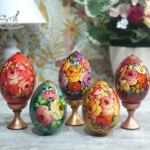 Cinq œufs en bois peints à la main avec des motifs floraux sur des supports dorés. Les œufs sont décorés de fleurs rouges, roses, jaunes, vertes et violettes. Les supports sont de couleur or brillant.