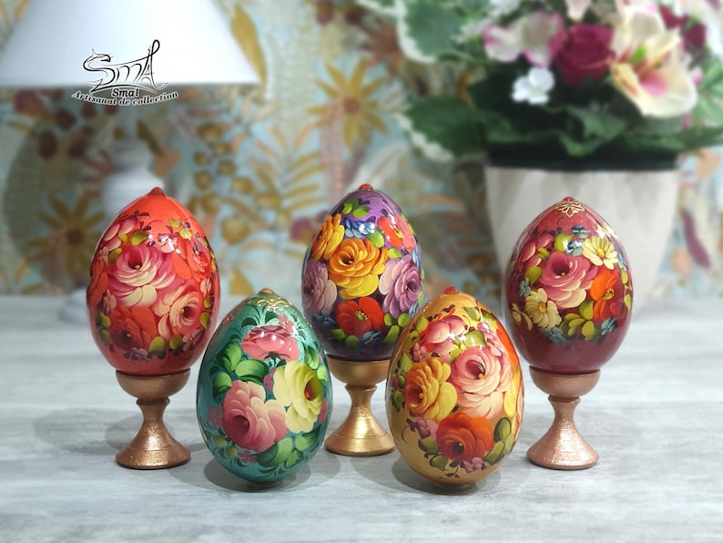 Cinq œufs en bois peints à la main avec des motifs floraux sur des supports dorés. Les œufs sont décorés de fleurs rouges, roses, jaunes, vertes et violettes. Les supports sont de couleur or brillant.