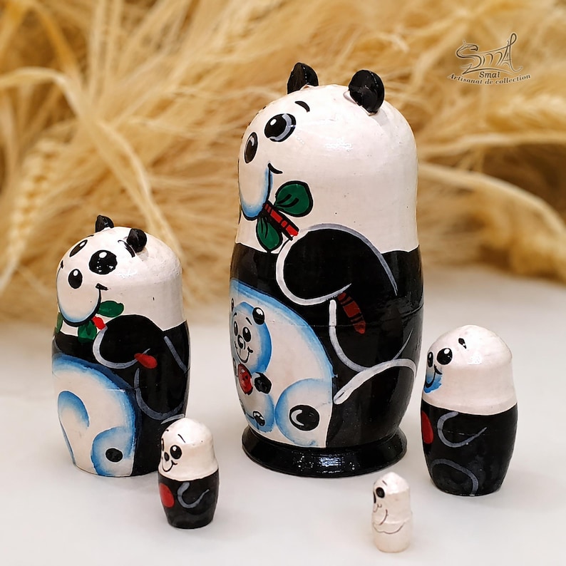Panda matryoshka nesting dolls displayed separately on a white tabletop, with the largest doll shown from a side angle. The softly blurred background features natural dried grasses, creating a warm, rustic product photography setting